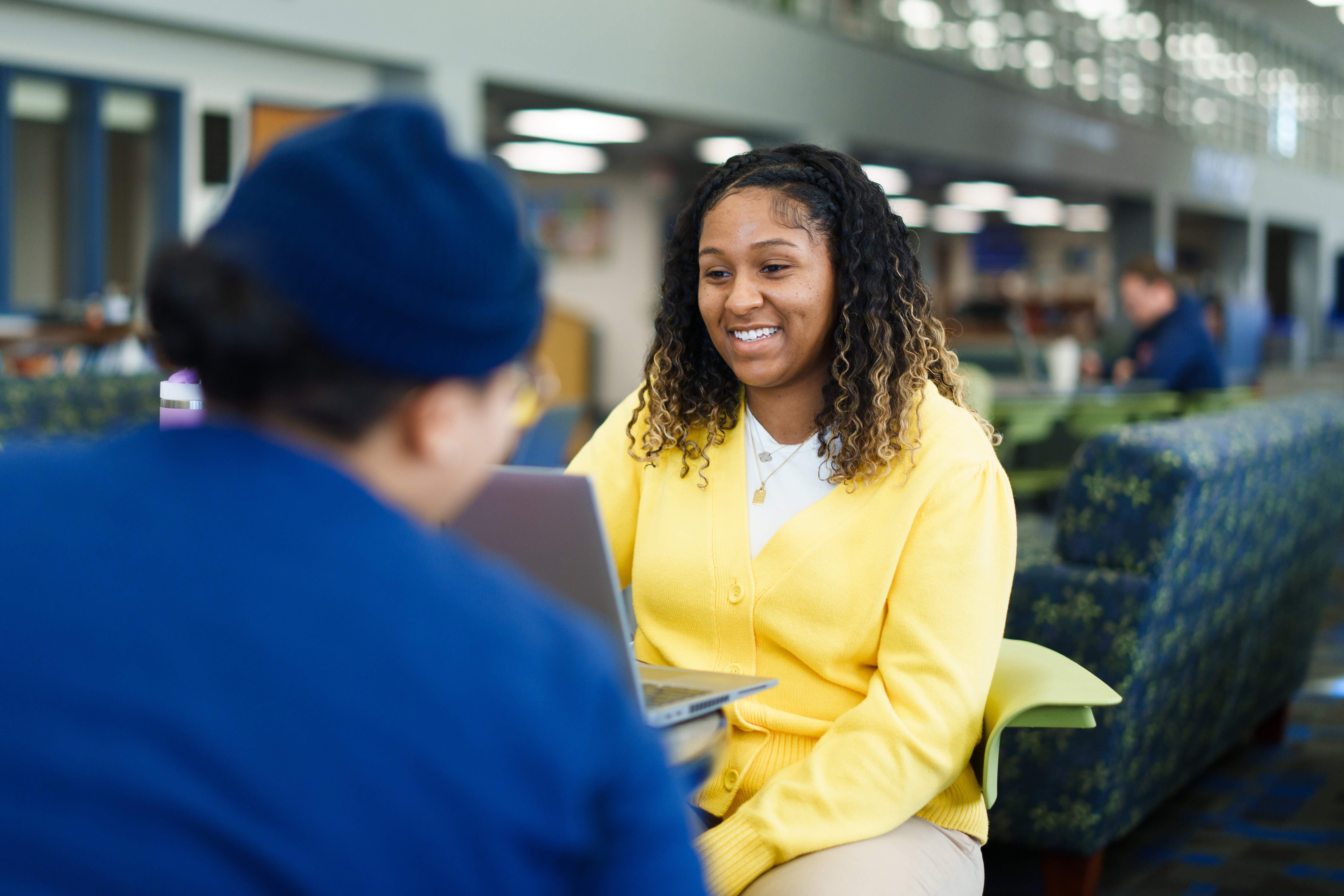 Two people sitting across from one another at a table; one is in focus, smiling and looking at laptop, while the other is blurred with their back turned to the camera